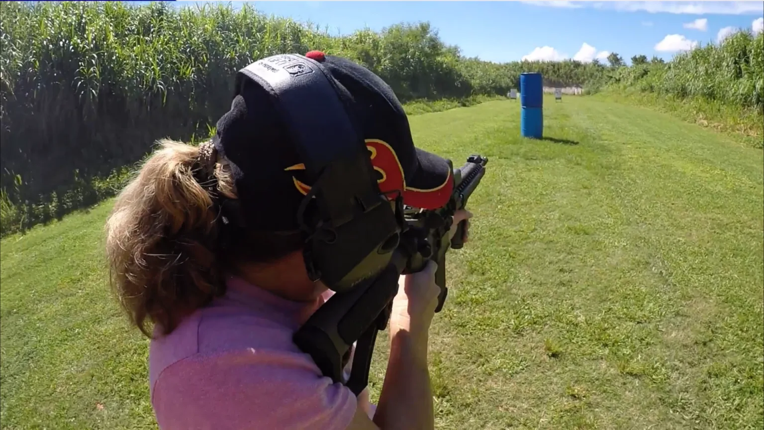 Woman shoulders a rifle beside a blue barrier on an outdoor range.