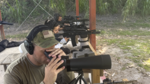 Shooter looks through a spotting scope while a rifle rests on support.