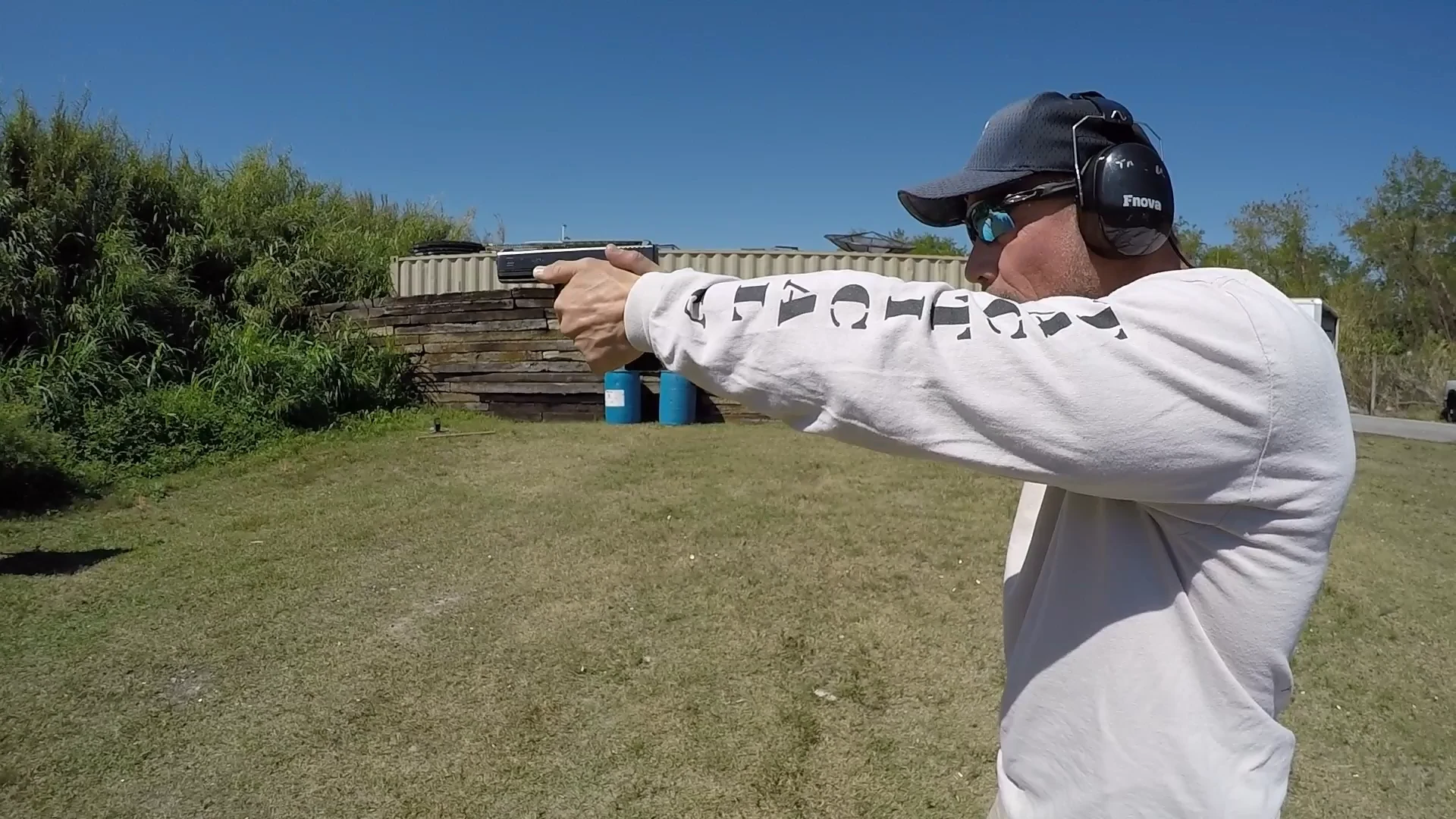 Stephen L. Cohen wearing a Tactical U shirt and hearing protection, aiming a pistol to the left on an outdoor range