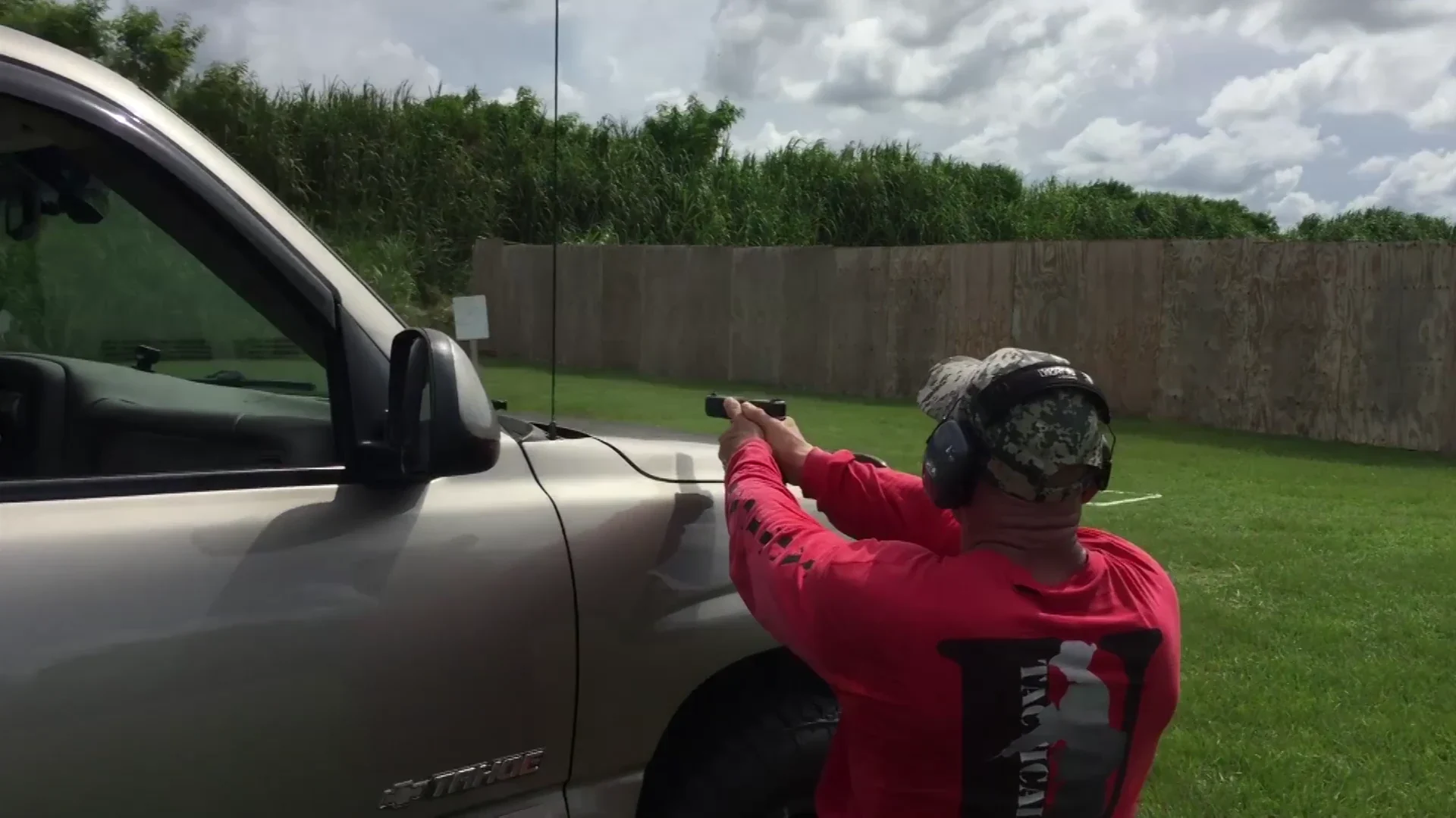 A participant in a red Tactical U shirt aiming a handgun beside a pickup truck on an outdoor range