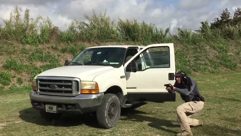 Student crouched at the front of a pickup truck aiming a pistol