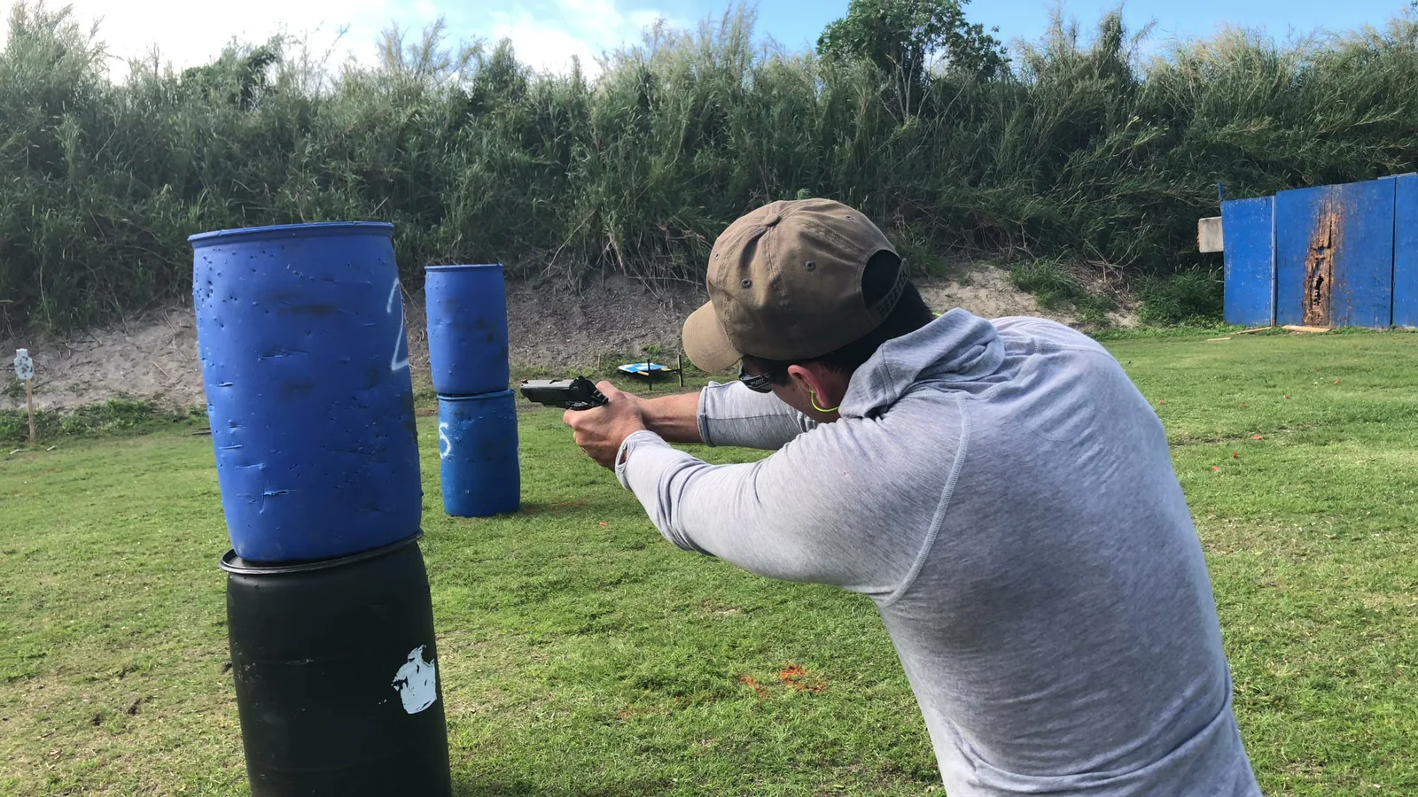 Shooter in a cap and ear protection firing a pistol from beside a stack of blue barrels
