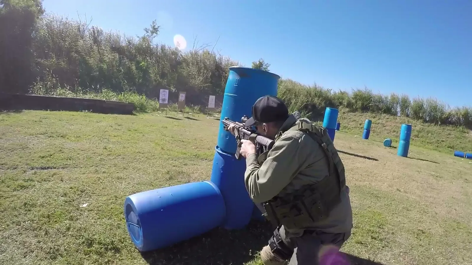 Rifle shooter kneeling and firing around a blue barrel used as cover on an outdoor range