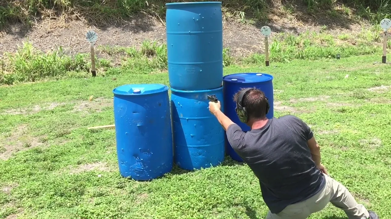 Shooter leaning around stacked blue barrels to engage a target on an outdoor range