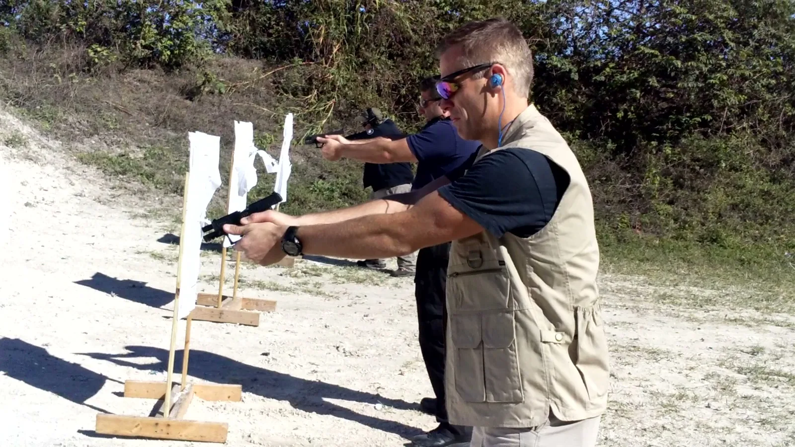 Instructor in a vest standing beside a student aiming a pistol at targets on a sandy outdoor range