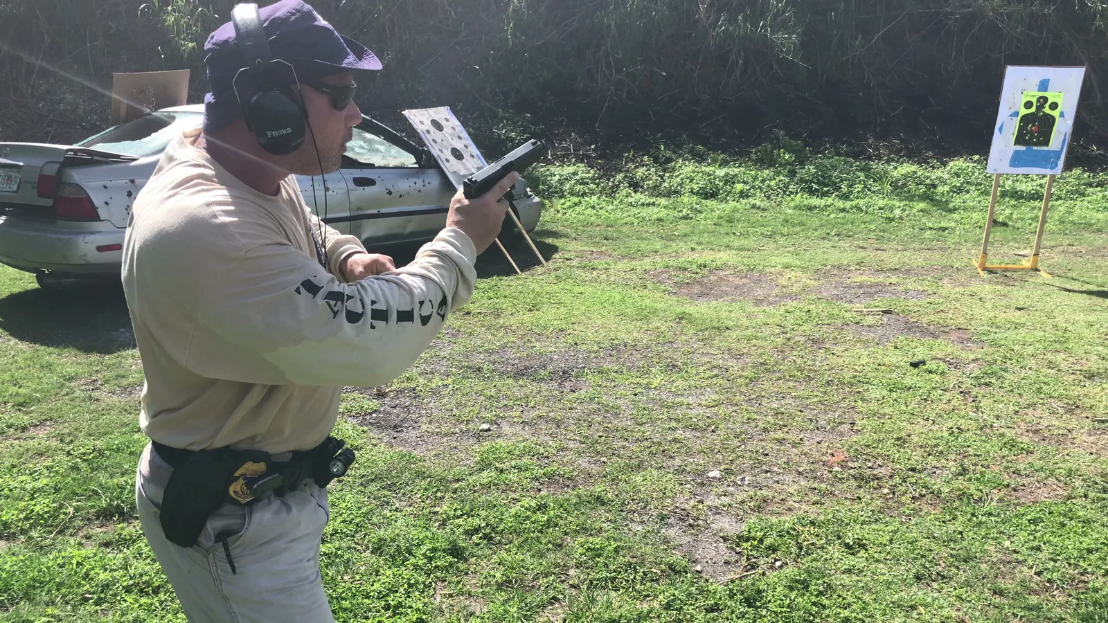 Tactical U instructor in ear protection working the slide of a pistol beside a parked car on an outdoor range