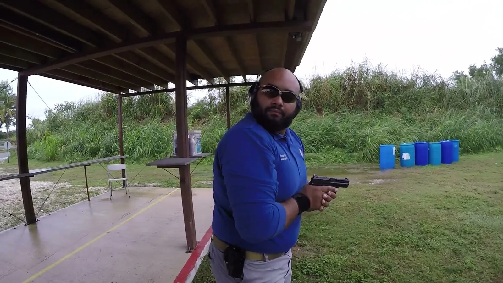 Shooter in a blue shirt holding a pistol low and safe at a covered outdoor firing line, wearing eye and ear protection
