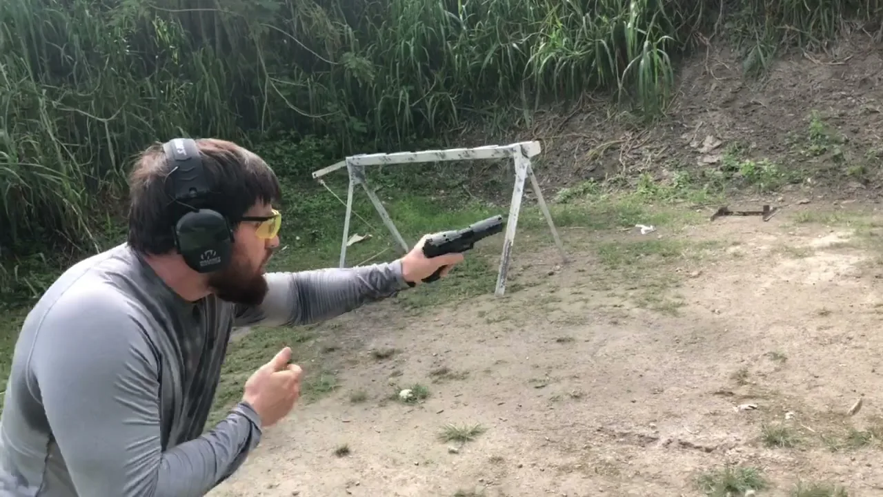 Shooter in ear protection firing a pistol one-handed beside a wooden barricade, support hand held back