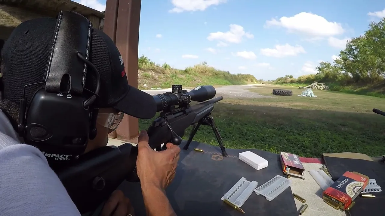 Scoped precision rifle on a bipod with ammunition laid out at the firing line, seen over the shooter’s shoulder