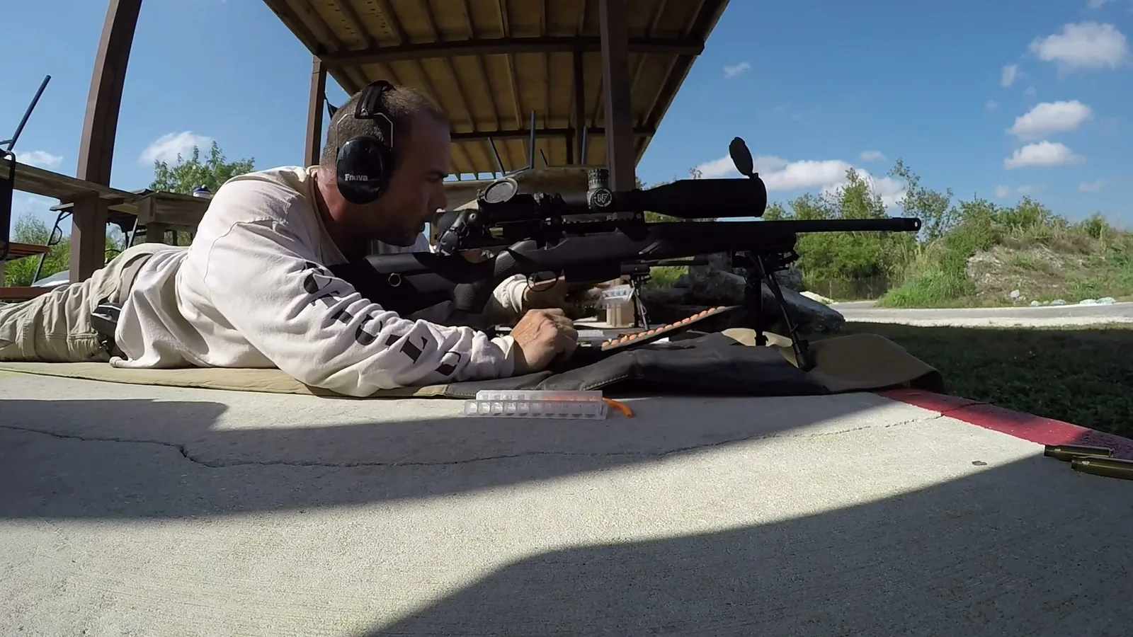 Shooter in the prone position behind a scoped precision rifle on a mat under the covered firing line