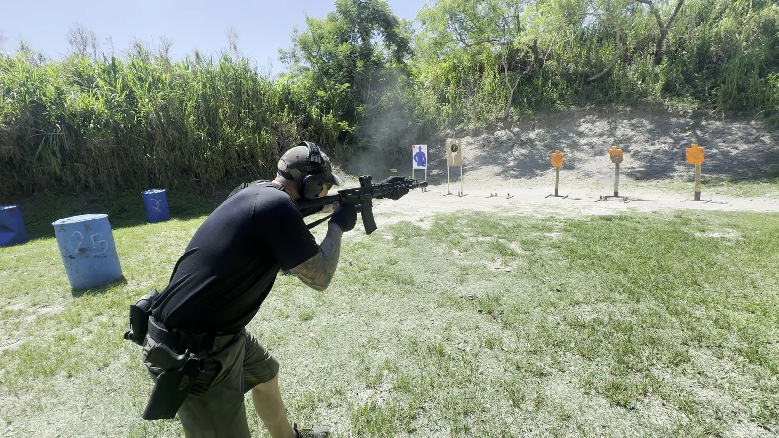 Rifle shooter moving between blue barrels while firing on an outdoor range