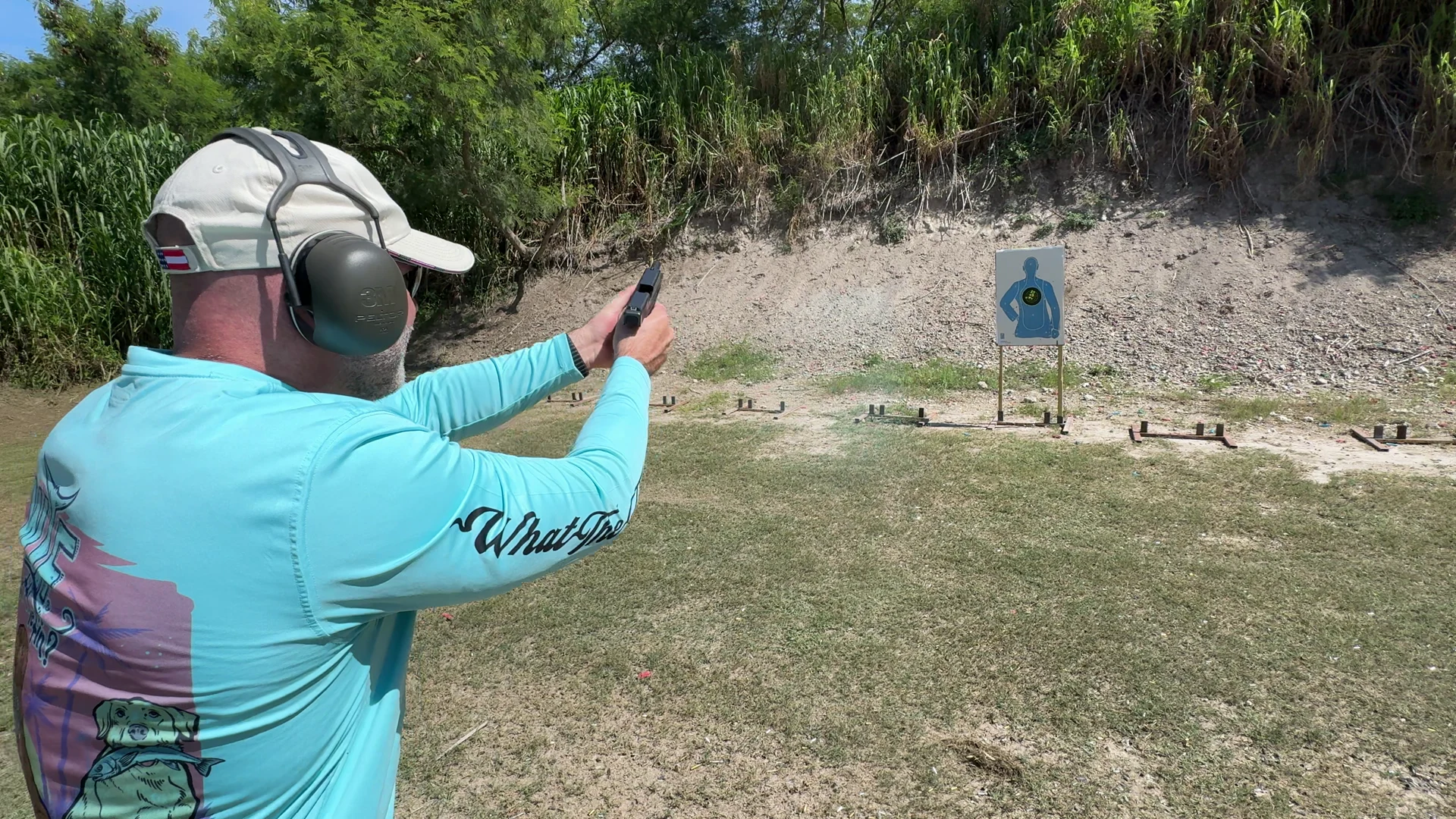 Over-the-shoulder view of a shooter aiming a pistol with both hands at a target on an outdoor range
