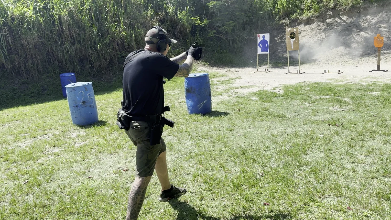 Student firing a pistol while moving between blue barrels toward targets on an outdoor range