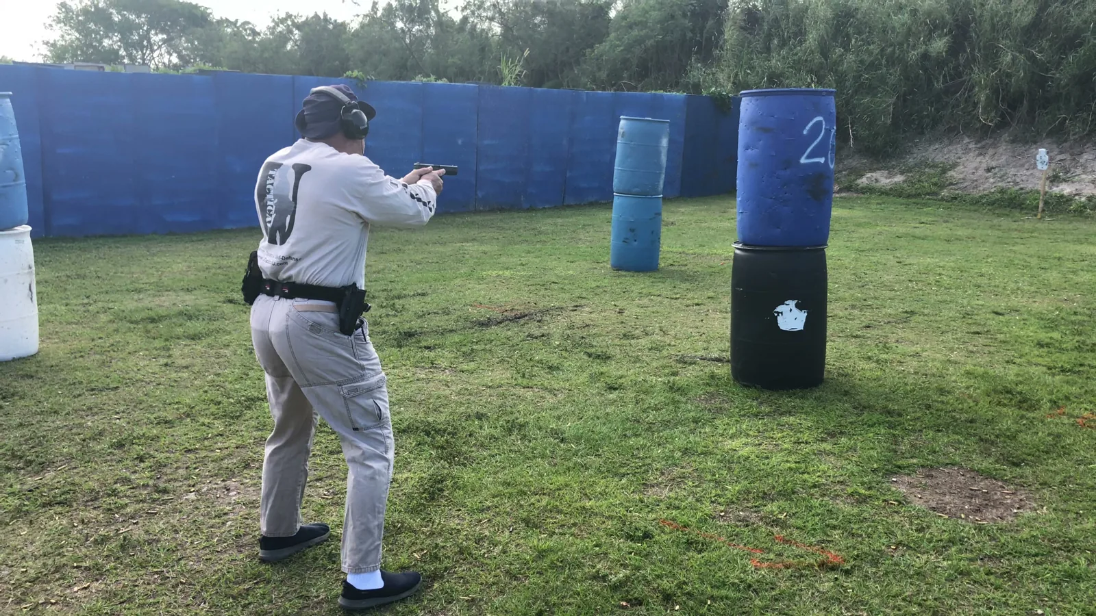 Tactical U student in a Tactical U shirt firing a pistol beside stacked barrels used as cover on an outdoor range