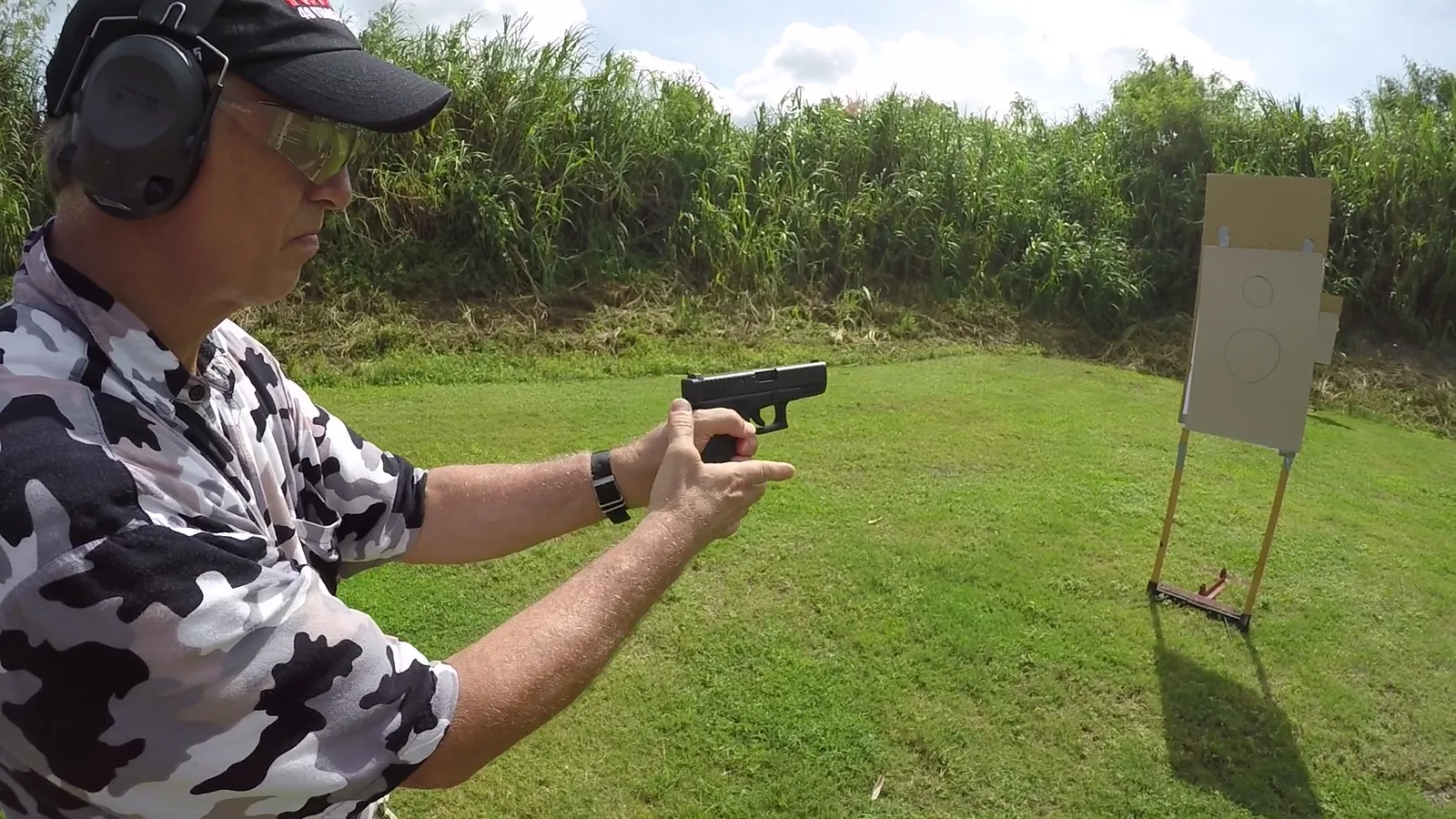 Student in a camouflage shirt and ear protection handling a pistol on an outdoor range