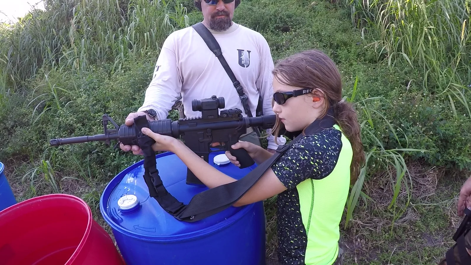 Young shooter in ear protection aiming a rifle supported on a blue barrel, with an instructor nearby